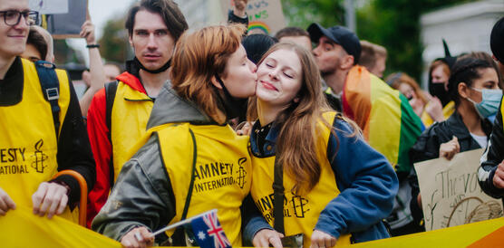 Das Foto zeigt eine Gruppe junger Menschen, die Leibchen mit dem Amnesty-Logo tragen. Sie stehen inmitten einer großen Menschenmenge. In der Bildmitte küsste eine junge Frau eine andere junge Frau auf die rechte Wange.
