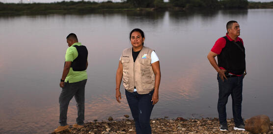 Das Bild zeigt drei Personen, die am Ufer eines Flusses stehen