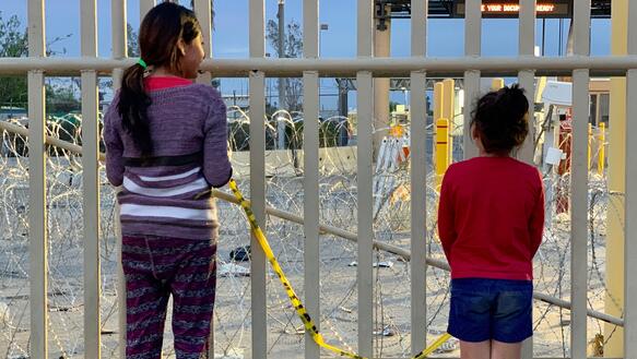 Ein älteres und ein jüngeres Mädchen stehen vor einem Zaun und blicken durch die Gitterstäbe.