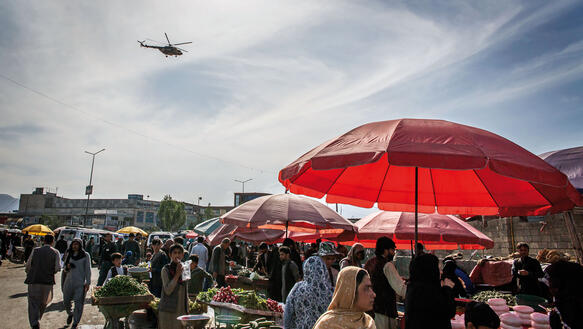 Ein Marktplatz mit Menschen, im Vordergrund ein Sonnenschirm, am Himmel ein Hubschrauber