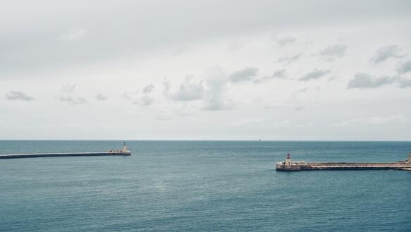 Foto vom Meer mit grauem Himmel, links und rechts historische Türmchen und Mauern eines Hafenbeckens
