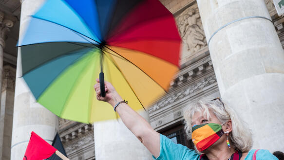 Eine Frau streckt einen Regenschirm in den LGBTI-Farben in die Höhe. Sie trägt eine Gesichtsmaske und Ohrschmuck, beides auch in den LGBTI-Farben.