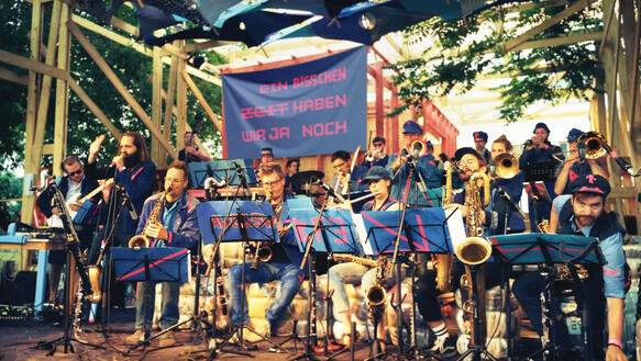 Eine Jazzkapelle mit vielen Musiker*innen auf einer Open-Air-Bühne, hinter ihnen hängt ein Plakat mit der Aufschrift "Ein bisschen Zeit haben wir ja noch".